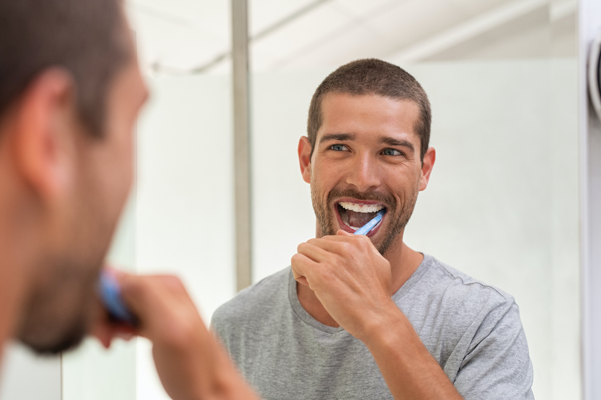 A man in a gray t-shirt brushes his teeth while looking at himself in the bathroom mirror